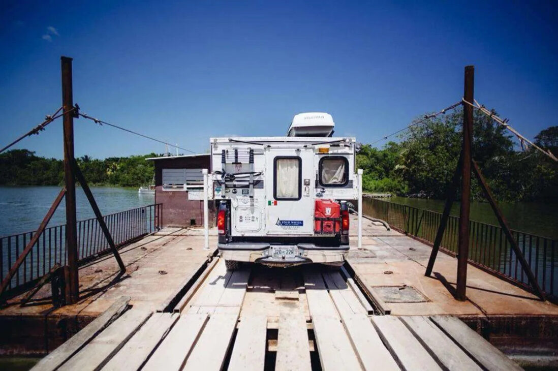 Traseira de uma Ford F-250 com camper posicionada sobre uma balsa cercada por água e vegetação em Belize.
