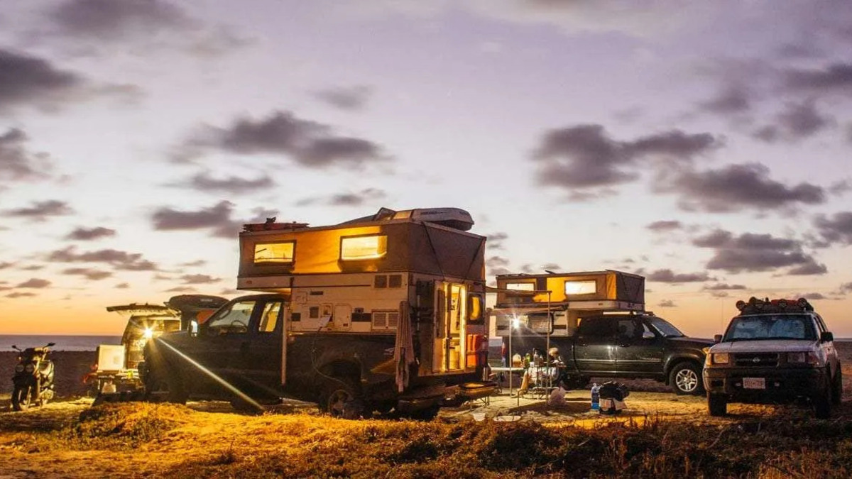 Ford F-250 com camper estacionada em acampamento à beira-mar durante o entardecer, ao lado de outros veículos adaptados.