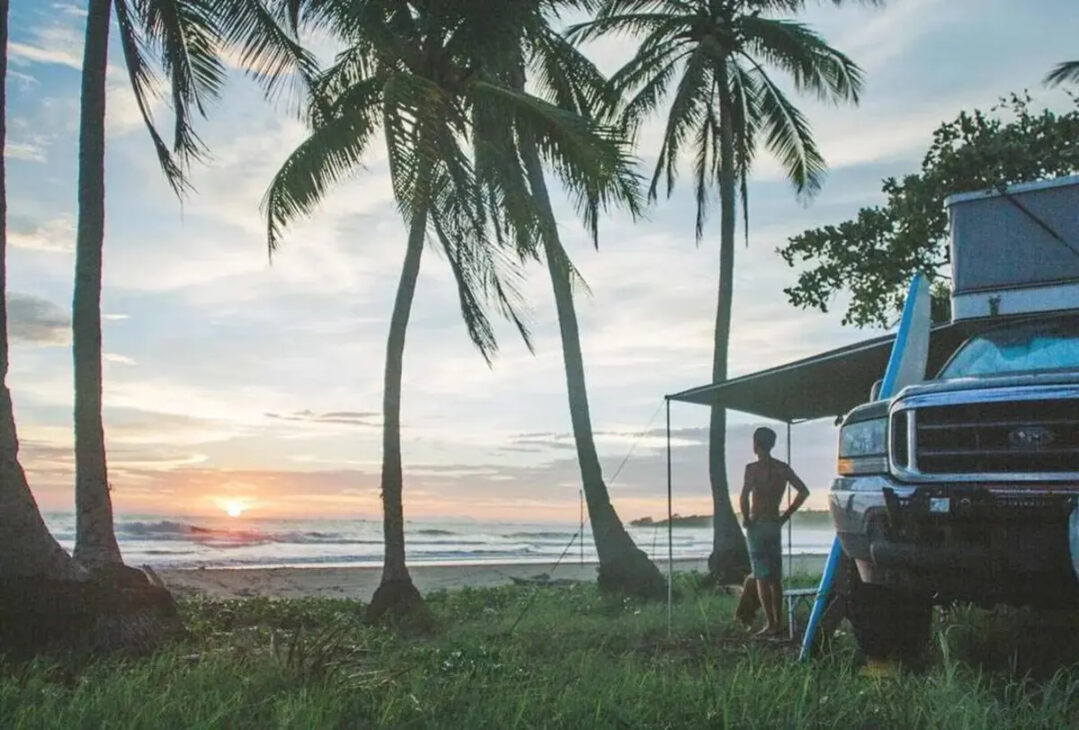 Ford F-250 com camper estacionada entre coqueiros diante do mar, enquanto um viajante observa o pôr do sol.