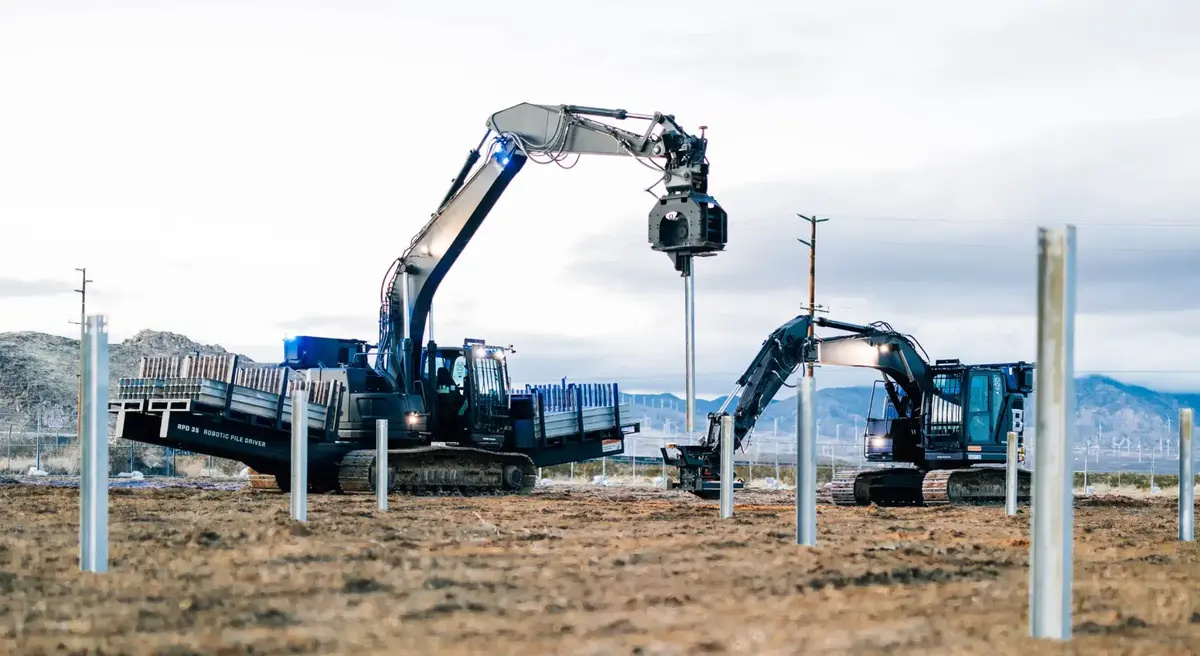 Duas máquinas autônomas transportam e cravam estacas metálicas durante a construção de uma usina solar.