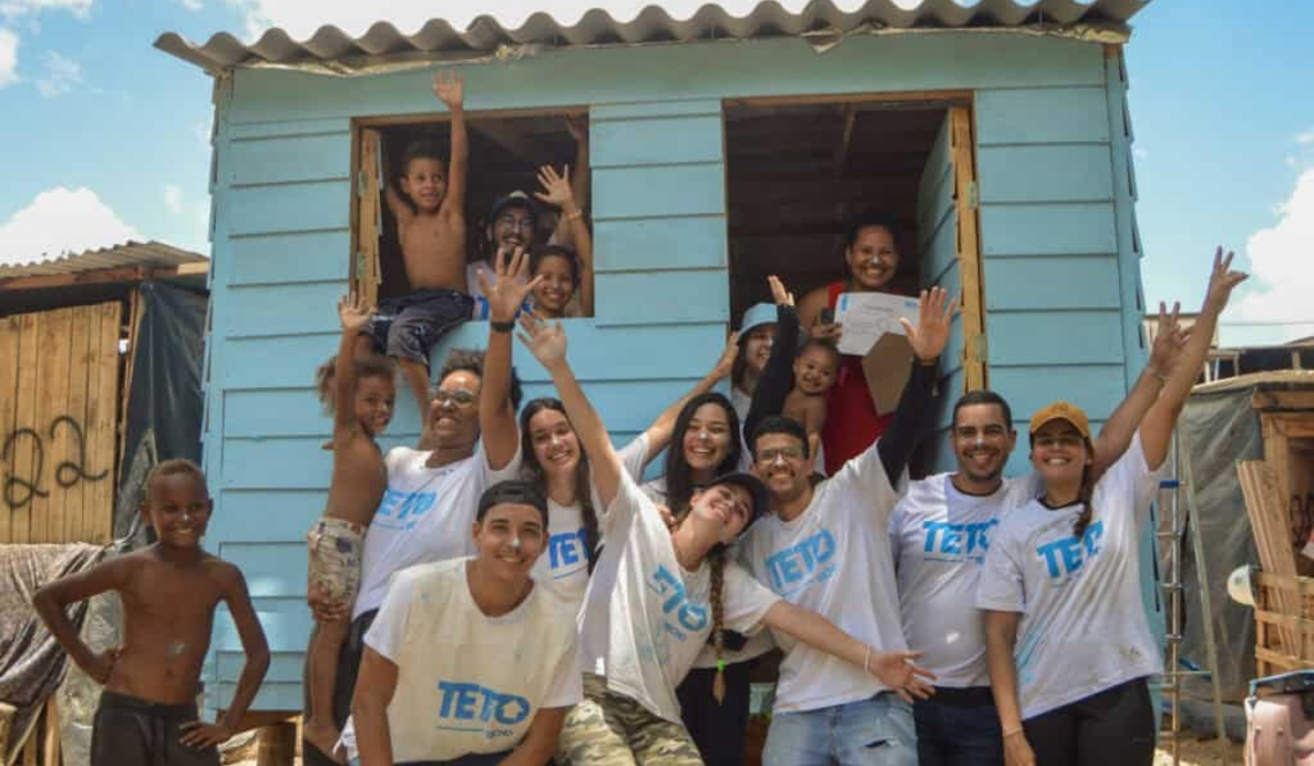 Moradores e voluntários da TETO diante de uma moradia emergencial construída com painéis pré-fabricados de madeira, modelo utilizado pela organização em seus mutirões habitacionais. - Foto de Arquivo: TETO Brasil/Reprodução