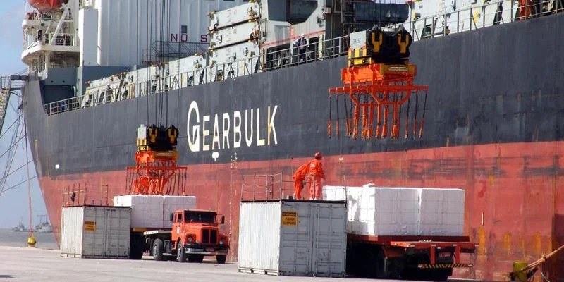 Fardos de celulose sendo embarcados em navio graneleiro no cais, com caminhões ao lado; imagem ilustrativa