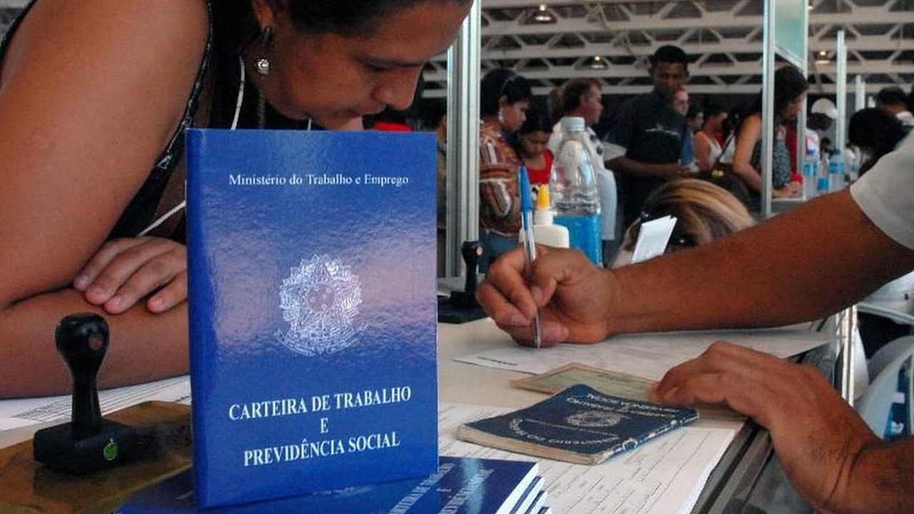 Carteira de Trabalho e Previdência Social azul sendo carimbada sobre um balcão de atendimento, com pessoas aguardando ao fundo