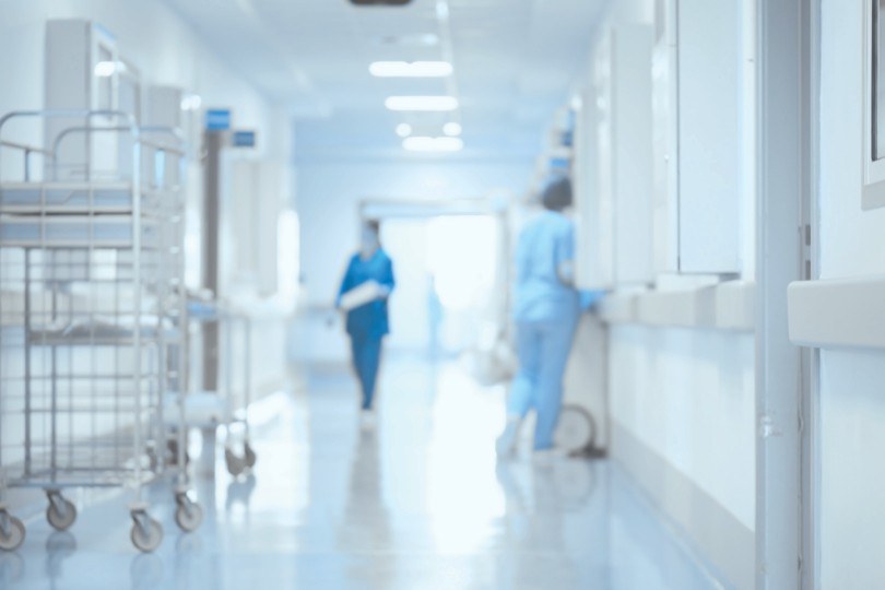 Corredor de hospital com carrinhos de material e equipe de enfermagem ao fundo; imagem ilustrativa