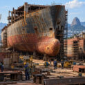 Trabalhadores e escavadeira industrial realizando desmantelamento de navio e separação de aço naval para reciclagem.