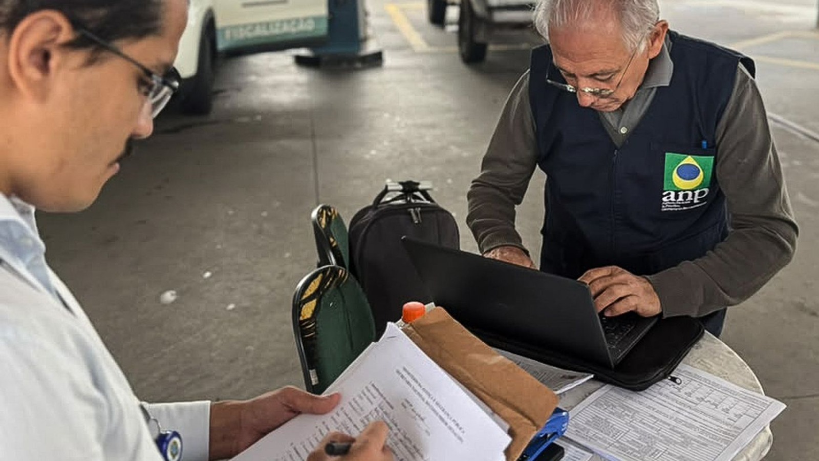 Fiscal de colete da ANP digita em notebook dentro de posto de combustível, ao lado de servidor que preenche formulário.