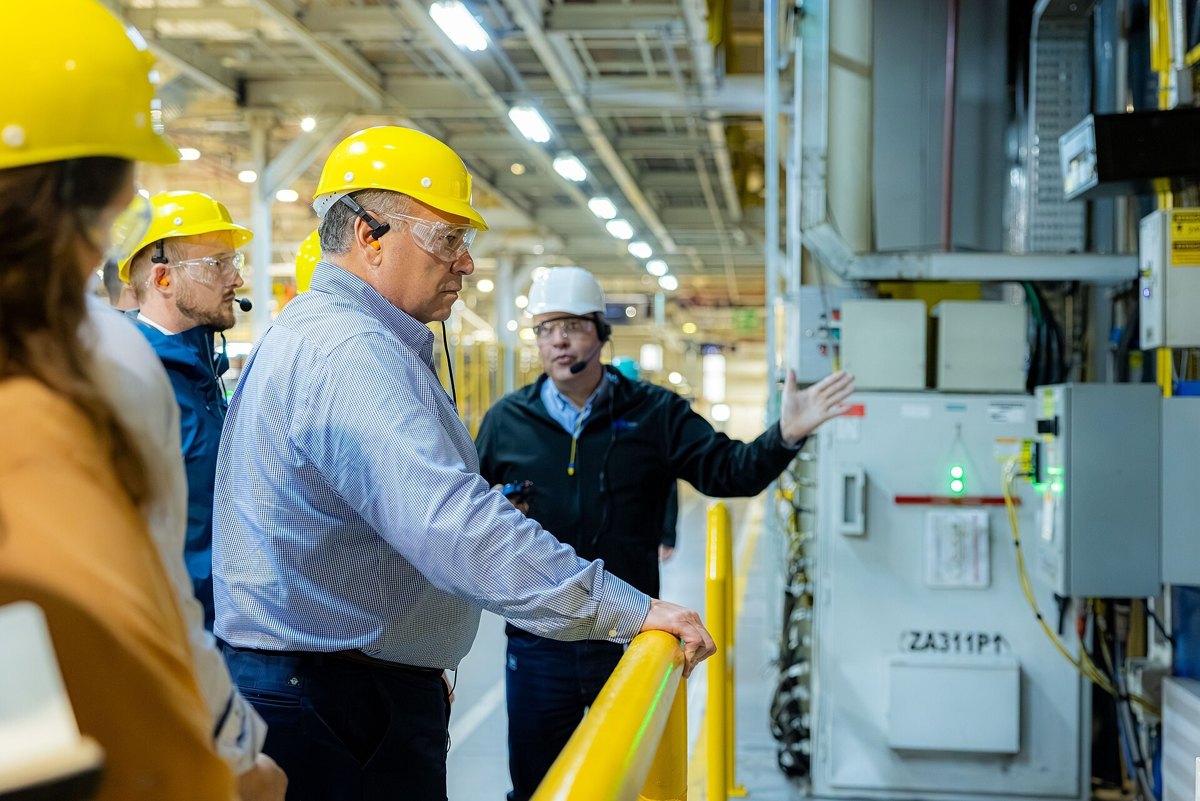Visitantes de capacete amarelo e óculos de proteção ouvem explicação ao lado de painel elétrico dentro de uma fábrica da General Motors