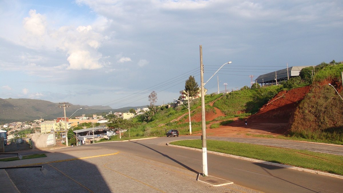 Avenida de Ouro Branco, em Minas Gerais, com morro de terra vermelha, postes e casas ao fundo sob céu azul