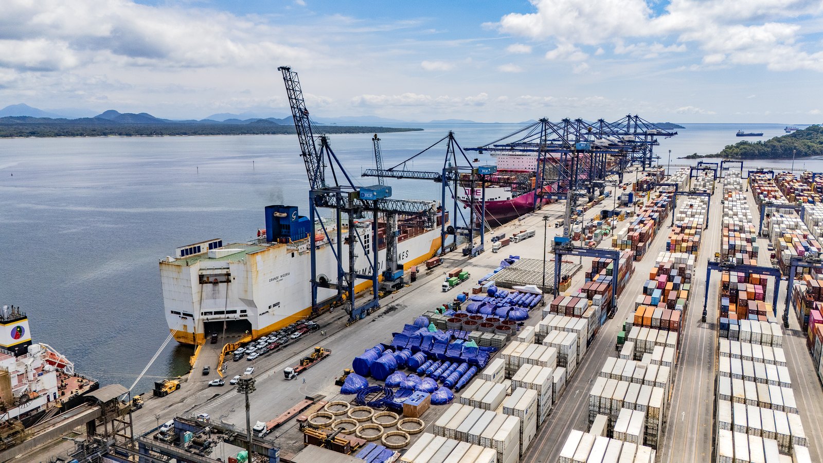 Vista aérea de um cais com guindastes de contêineres azuis, navios atracados e um pátio cheio de contêineres coloridos à beira da baía.