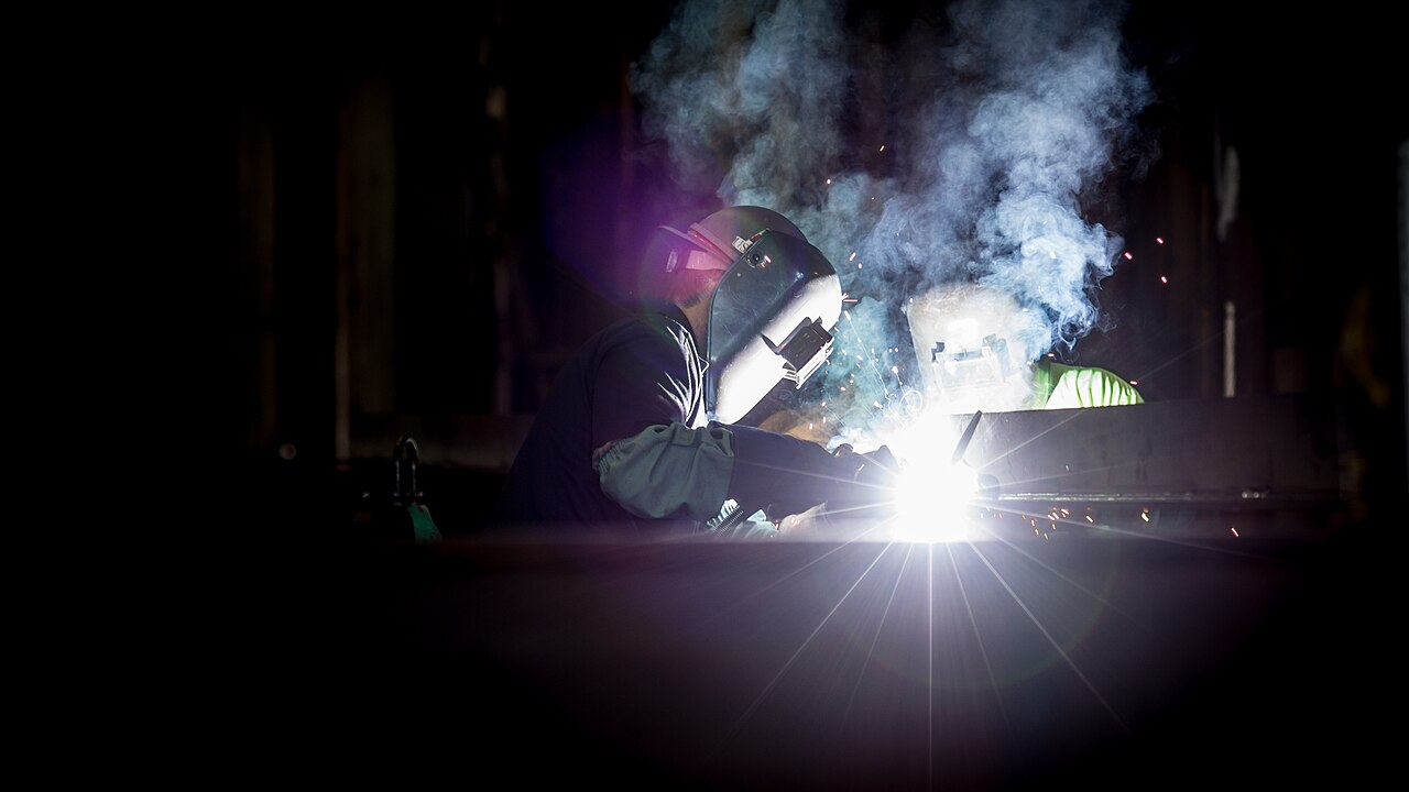 Dois profissionais usando máscaras de proteção soldam peças de aço em um estaleiro industrial